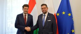 Przed białą ścianą stoją wiceminister Szeptycki i mężczyzna w ciemnogranatowym garniturze, podają sobie ręce, za nimi flagi Polski, Węgier i UE, po lewej zasłonięte firankami okno.