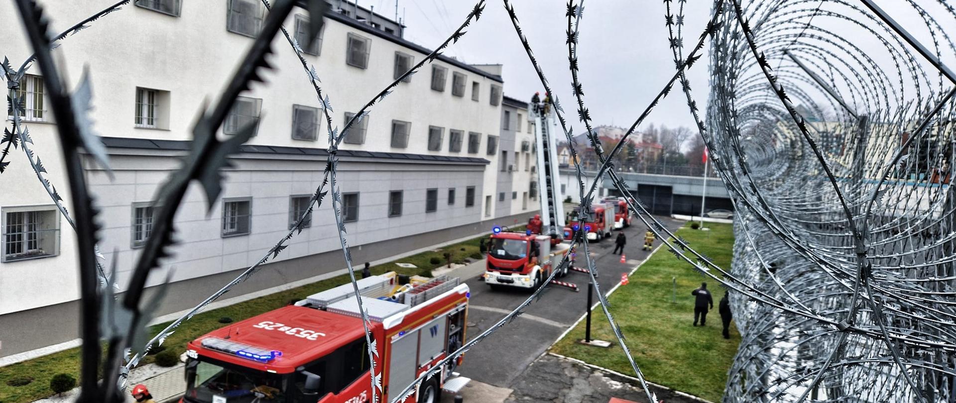 Na zdjęciu samochody bojowe Państwowej Straży Pożarnej na terenie Aresztu Śledczego widziane zza ogrodzenia z drutu kolczastego.