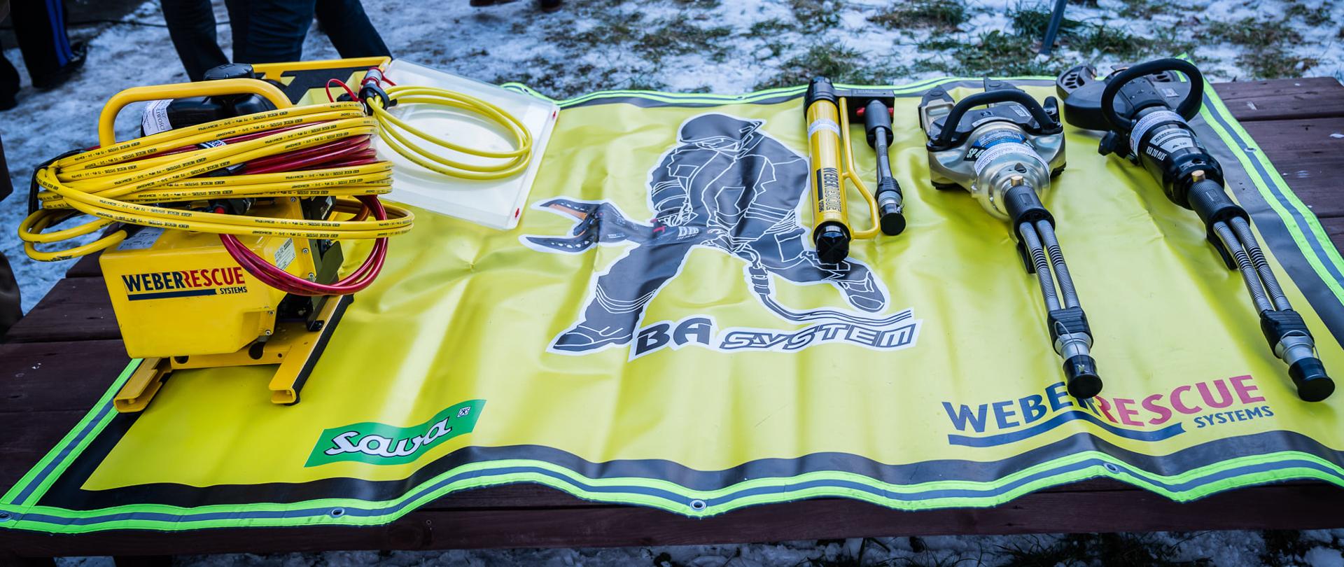 Zdjęcie przedstawia nowy zestaw hydrauliczny firmy WEBER Rescue System koloru żółtego. Zestaw leży na żółtej macie z wizerunkiem strażaka.