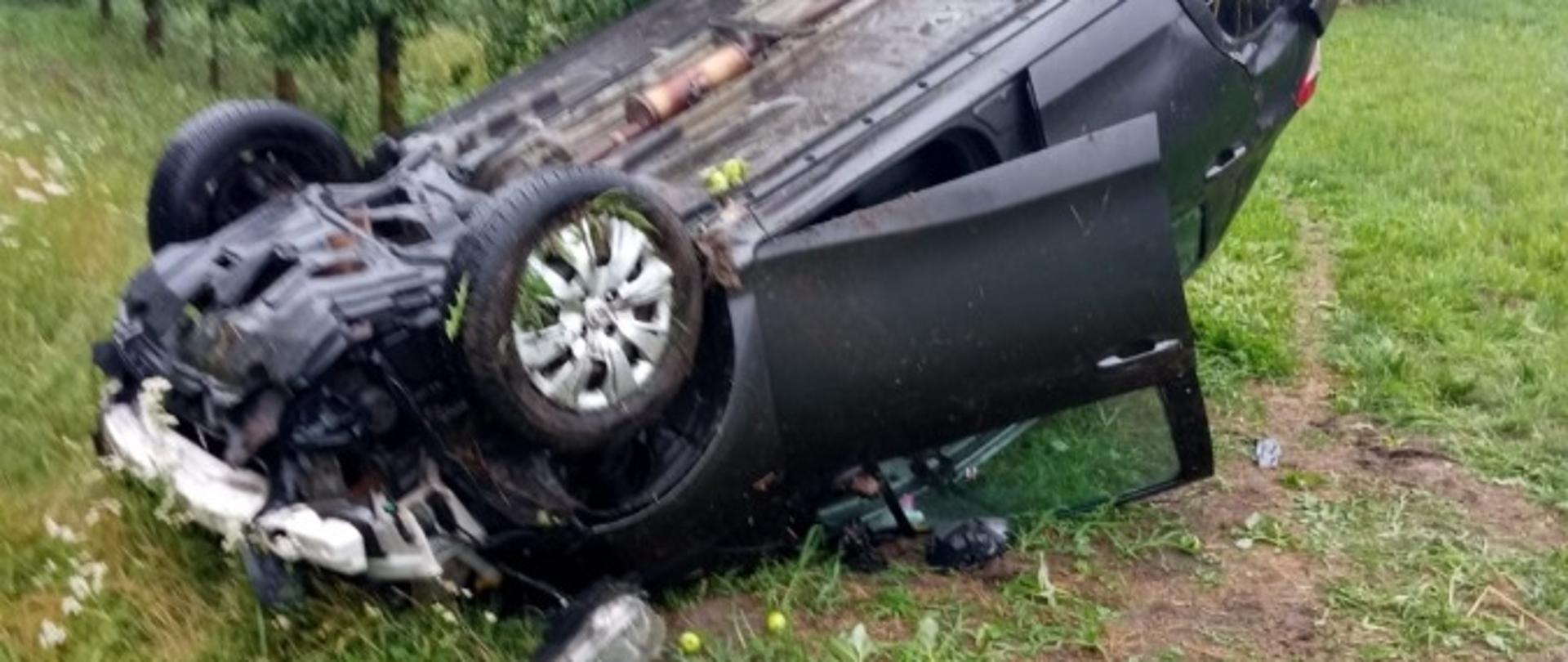 Na zdjęciu widoczny samochód osobowy koloru ciemnego leżący na dachu poza jezdnią. Na drugim planie sad owocowy.