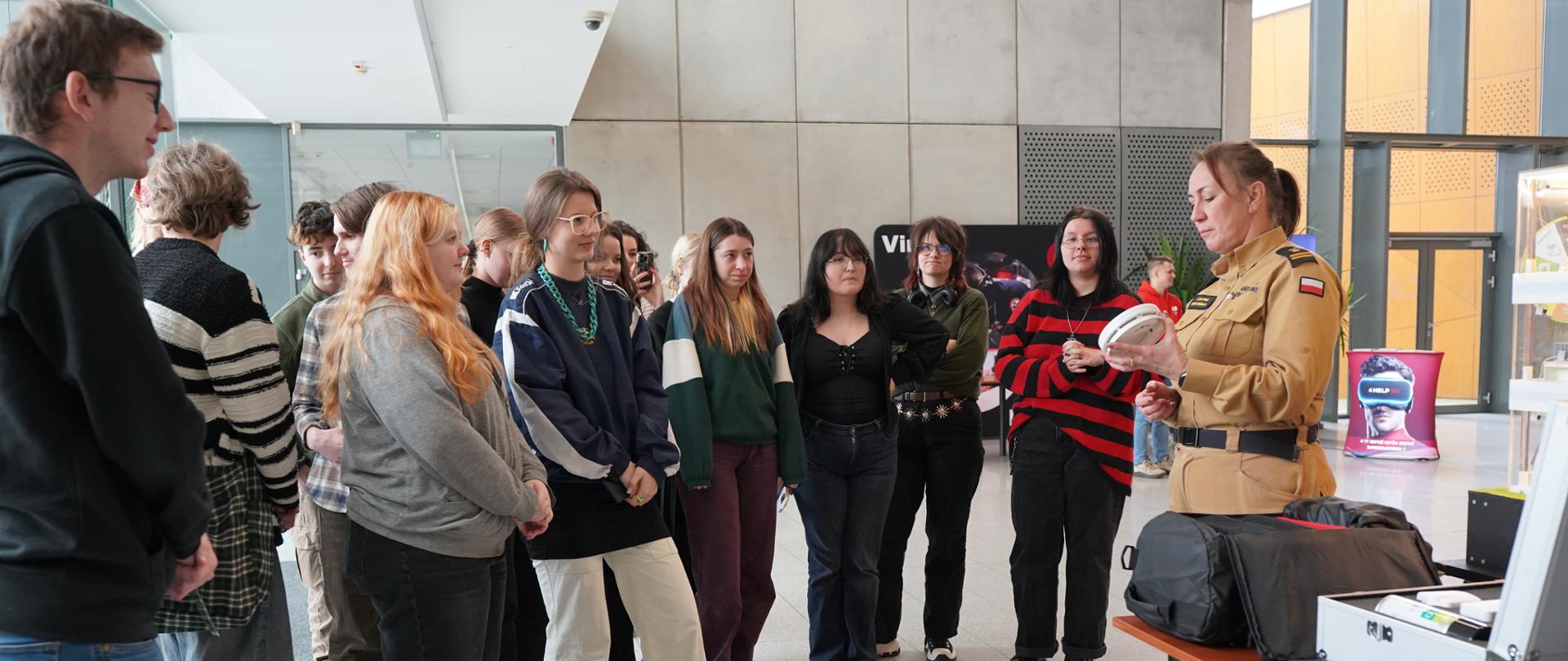 W jasnym holu grupa młodych osób stoi w półkolu i słucha kobiety strażaka w beżowym mundurze, która prezentuje czujkę. Na stoliku obok niej leżą torby i sprzęt techniczny. W tle widać przeszklone wejście oraz plakaty wystawowe.