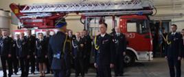 Zastępca Mazowieckiego Komendanta Wojewódzkiego PSP st. bryg. Piotr Gąska przyjmuje meldunek dowódcy uroczystości. Obaj stoją na tle grupy strażaków w mundurach wyjściowych stojących wzdłuż boku samochodu strażackiego z drabiną mechaniczną. Zdjęcie wykonane w pomieszczeniu garażowym. 