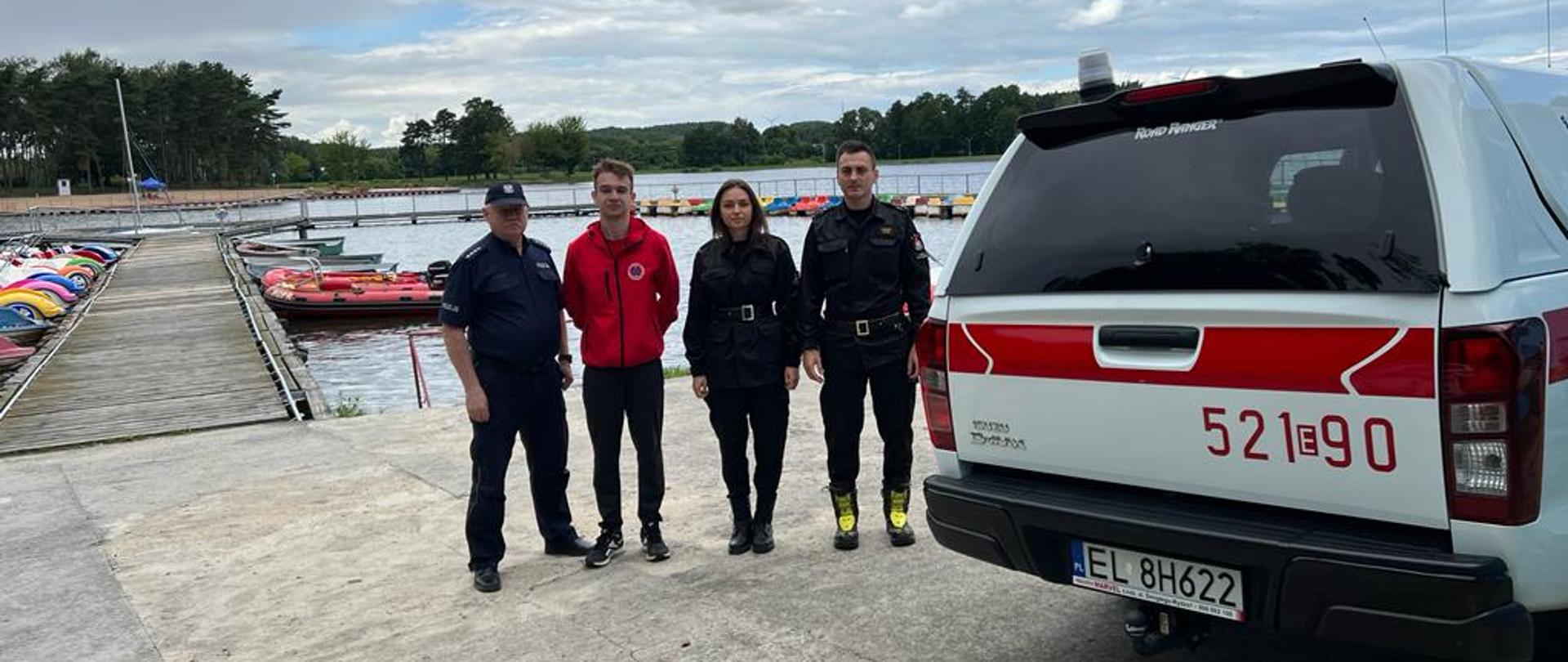 Cztery osoby, 3 mężczyzn i 1 kobieta, stoją na przystani w jednym szeregu. Kolejno od lewej: policjant z mundurze, ratownik WOPR, strażaczka i strażak w umundurowaniu. Po prawej stronie stoi biało- czerwony samochód terenowy ISUZU należący do straży pożarnej. Za nimi widać zalew oraz przystań z rowerkami wodnymi oraz kąpielisko.