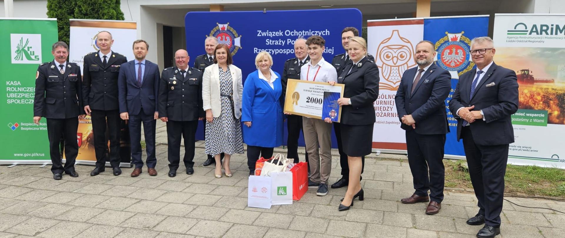Przed budynkiem na tle banerów organizatorów stoi grupa kobiet i mężczyzn, część z nich w mundurach straży pożarnej. W środku stoi młody mężczyzna z symbolicznym czekiem na 2000 złotych i dyplomem. Przed nim stoją torby z logotypami organizatorów turnieju.