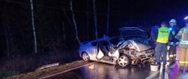 Rozbity samochód osobowy hiundai koloru srebrnego stoi częściowo na poboczu i na jednym pasie drogi. Obok stoi policjant i dwóch strażaków, 