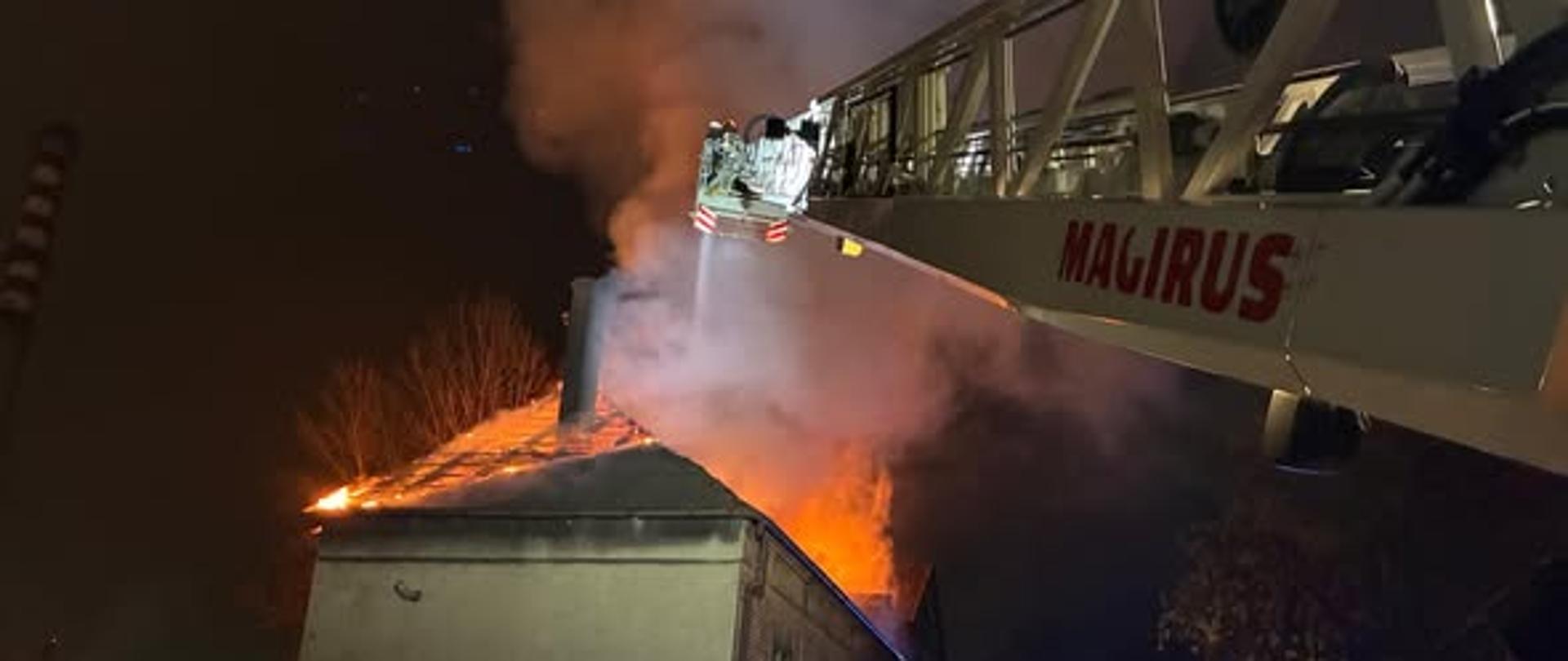 Na zdjęciu widoczny budynek z płonącym dachem. Do dachu sięga drabina strażacka. Z kosza drabiny podawana jest woda. Na ulicy przy budynku stoją wozy strażackie.