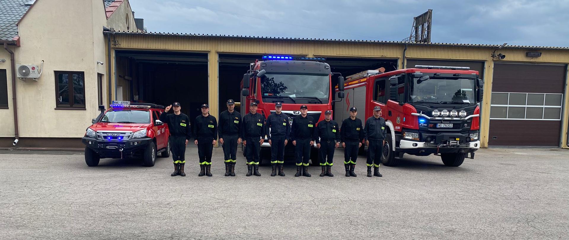 Zdjęcie przedstawia strażaków JRG Wołomin ustawionych przed pojazdami pożarniczymi. W autach włączone są sygnały świetlno-dzwiękowe. Dowódca pododziału oddaje honor po przez salut.