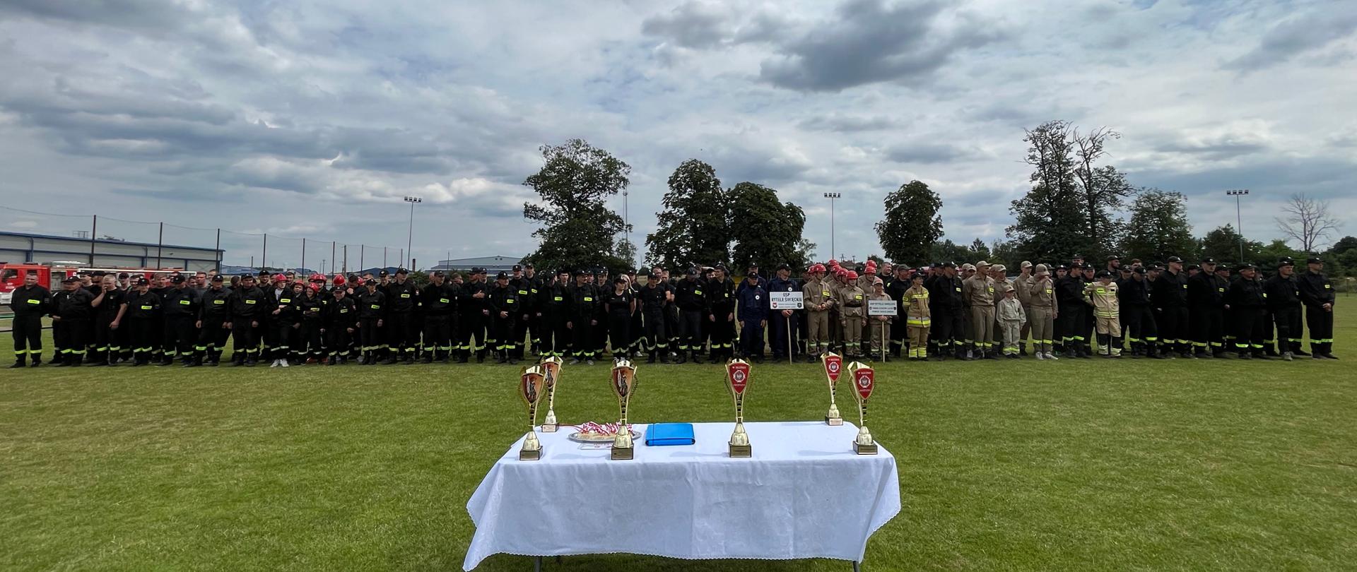 Międzygminne Zawody Sportowo – Pożarnicze jednostek OSP z terenu gmin Ceranów i Kosów - strażacy stojący na zbiórce i puchary stojące na stoliku.