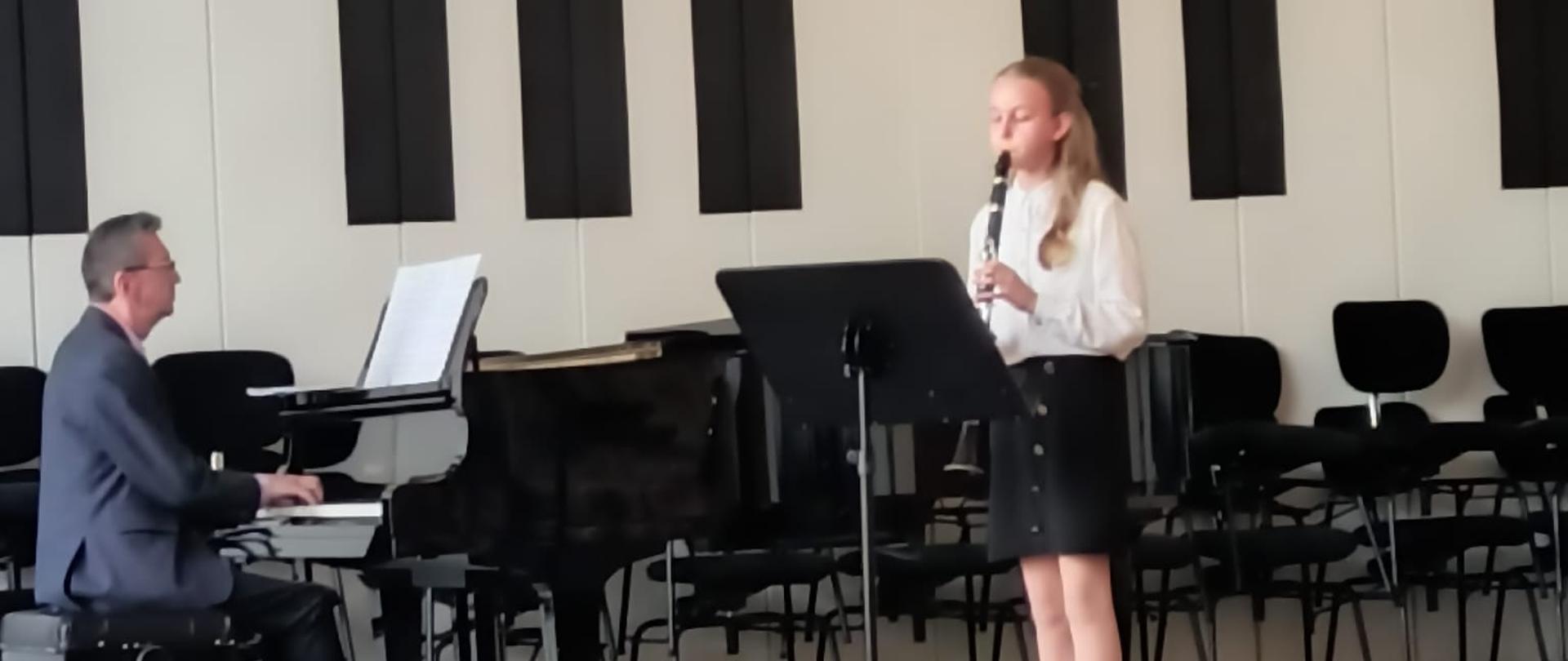 Dziewczynka gra na klarnecie, mężczyzna akompaniuje na fortepianie.