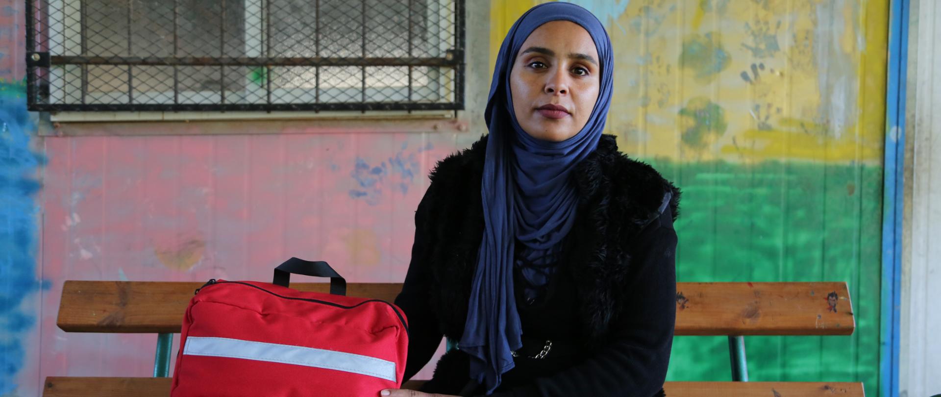 Kobieta w chuście z czerwonym plecakiem z napisem First Aid i logo PAH, Polska pomoc, Care