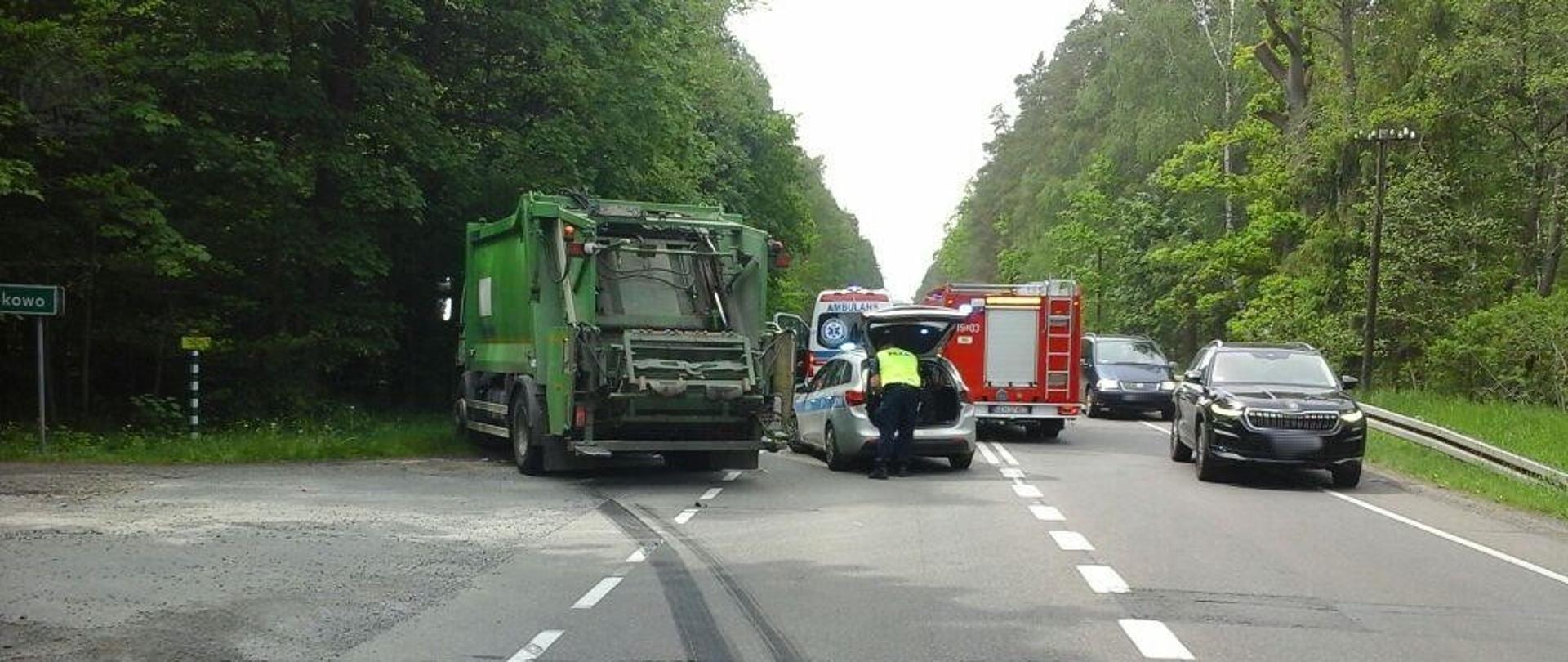 Śmieciarka uczestnicząca w wypadku. Widać ślady hamowania. Obok śmieciarki wóz strażacki, radiowóz policyjny oraz karetka. 