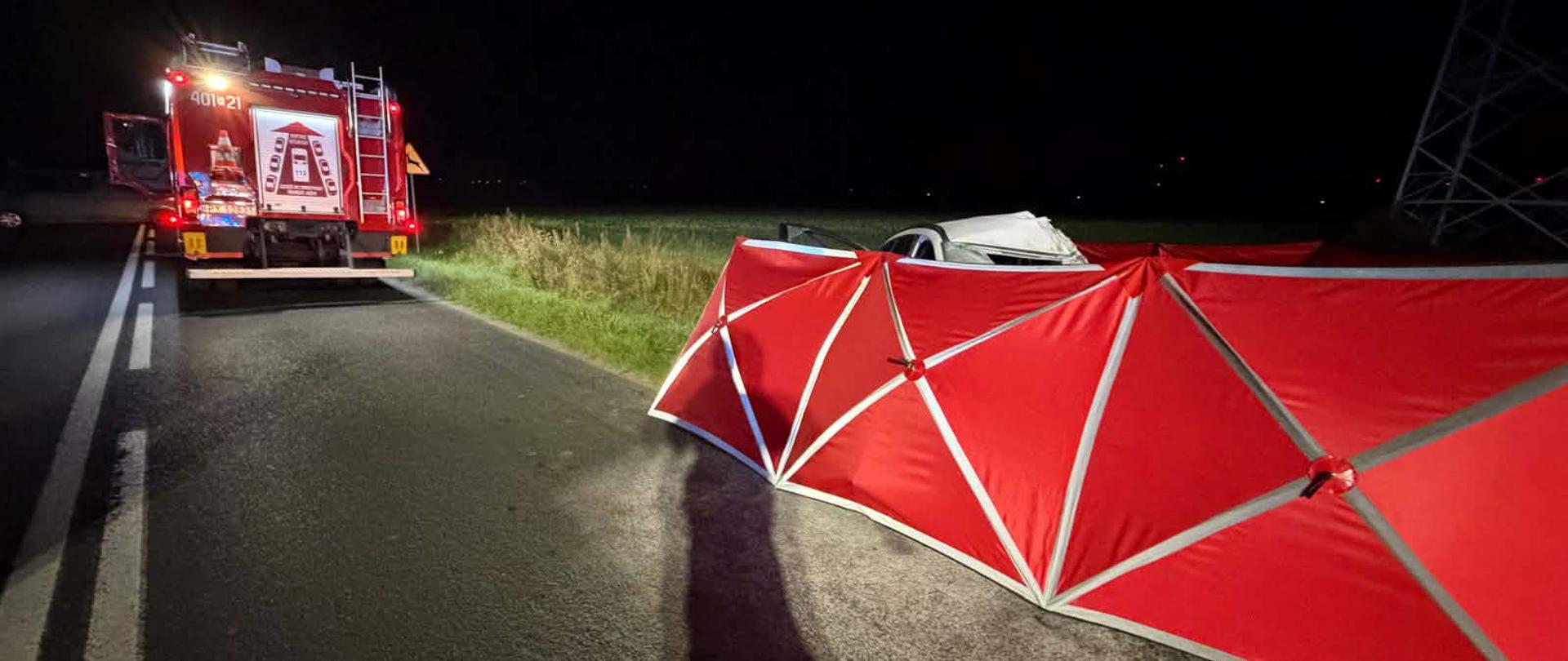 Zdjęcie przedstawia strażaków podczas wypadku drogowego śmiertelnego.
W tle samochody strażackie.
