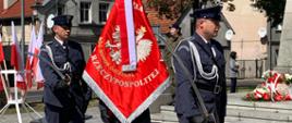 Rocznica wybuchu II WŚ. Zdjęcie przedstawia plac przed pomnikiem Żołnierza Polskiego. Na pierwszym planie poczet sztandarowy wystawiony przez Służbę Więzienną Zakładu Karnego w Rawiczu. Poczet maszeruje przed pomnik. Dowódca pocztu i asystujący w prawej ręce. W tle stojak z flagami biało-czerwonymi oraz zabudowania.