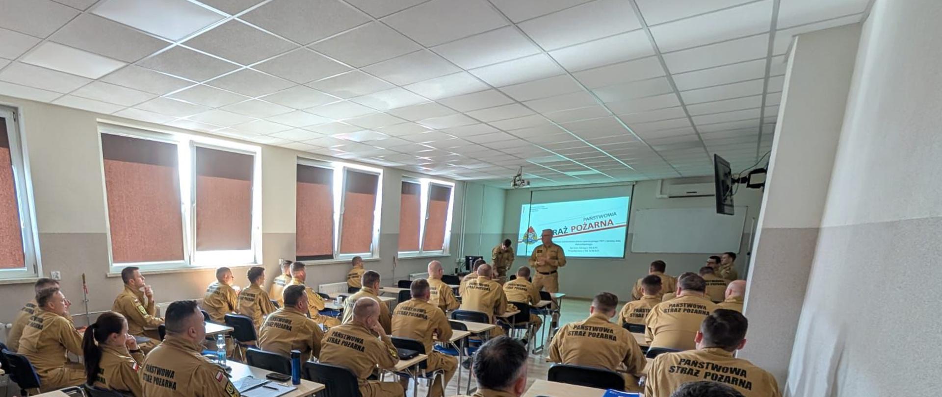 Zdjęcie przedstawia grupę strażaków podczas szkolenia teoretycznego w jasnej, nowoczesnej sali wykładowej. Funkcjonariusze siedzą w ławkach, zwróceni w stronę ekranu projekcyjnego, na którym widnieje napis „PAŃSTWOWA STRAŻ POŻARNA”.