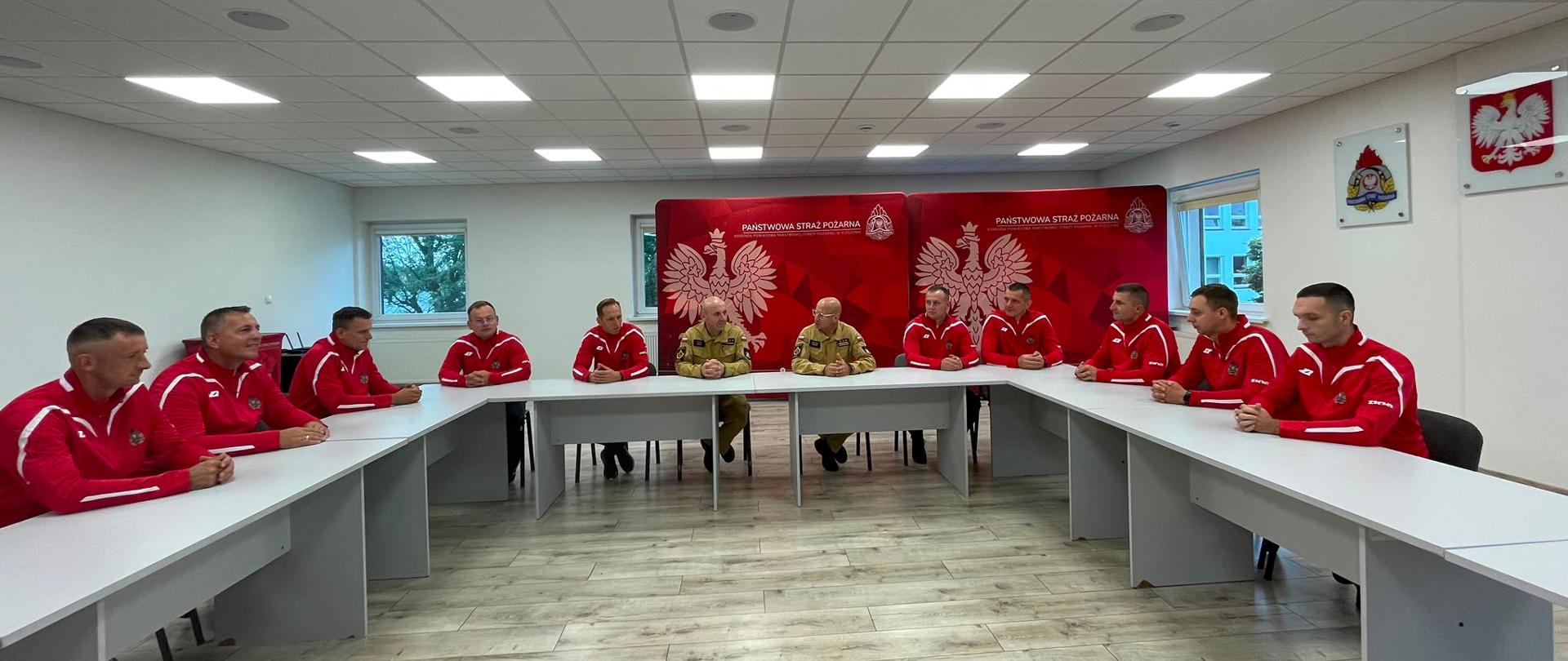 grupa osób w czerwonych dresach siedzący przy stole rozmawiają z przełożonym na sali konferencyjnej