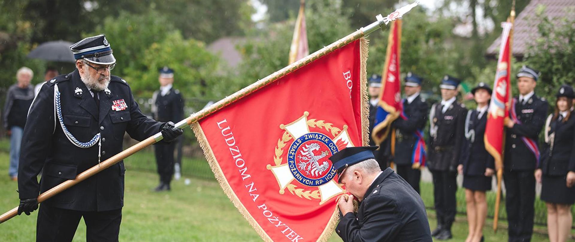 Strażak w mundurze całuje płat sztandaru jednostki OSP pochylony przez innego strażaka. Za nimi inni strażacy stoją w szyku ze swoimi sztandarami. Tło stanowi zieleń trawnika i drzew.