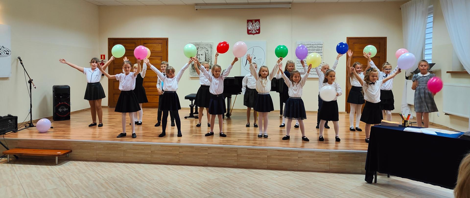 Zdjęcie przedstawia scenę sali koncertowej PSM w Grajewie, na której stoją uczniowie klas pierwszych ubrani na galowo. Dzieci trzymają nad głowami kolorowe baloniki. 