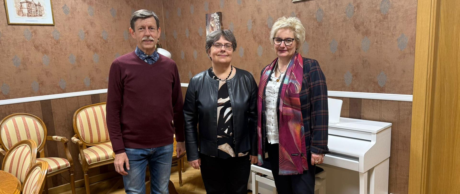 Trzy osoby stoją w eleganckim pomieszczeniu z drewnianą podłogą, brązowymi ścianami i klasycznymi krzesłami. Po lewej stronie stoi mężczyzna w bordowym swetrze i dżinsach, pośrodku kobieta w czarnej kurtce i czarnych spodniach, a po prawej kobieta w czarnych spodniach i marynarce w kratę z kolorową apaszką. W tle widoczny jest biały pianino oraz obrazy na ścianie.