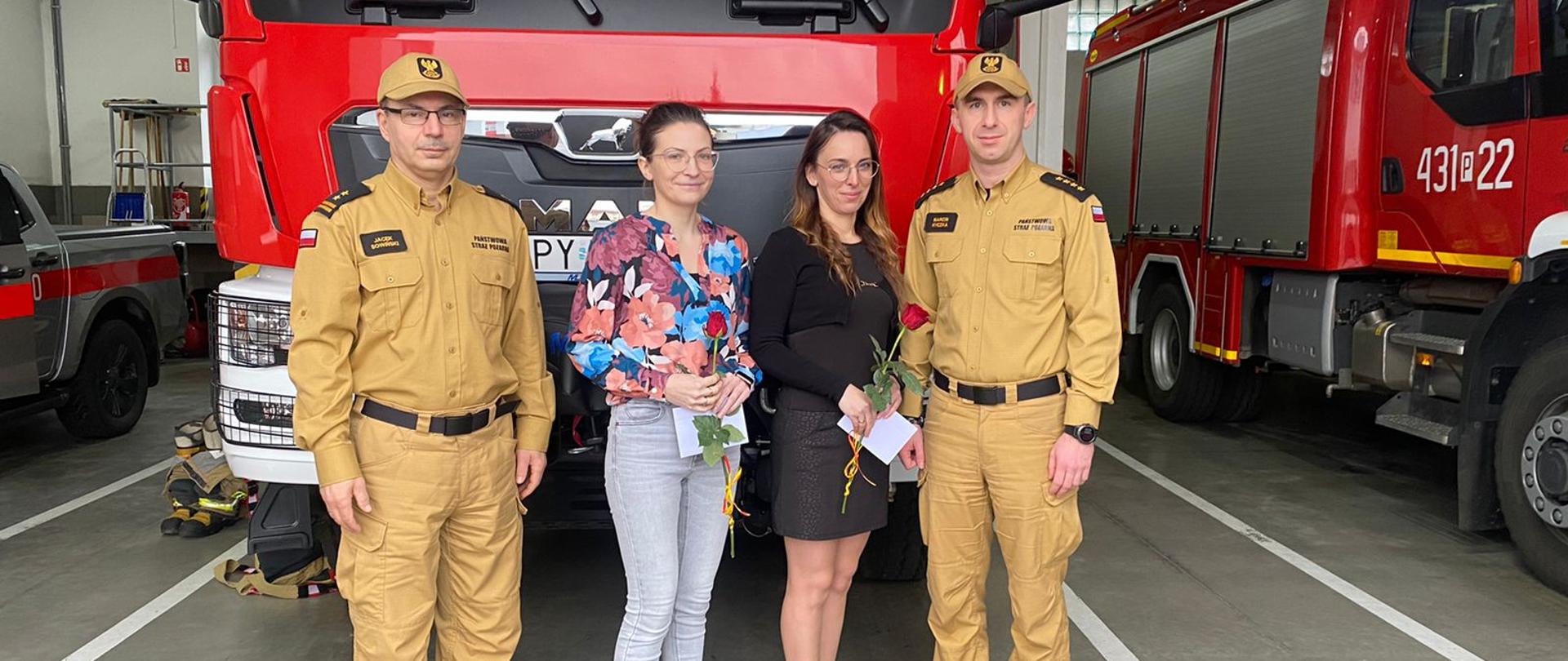 Komendantów oraz dwie kobity (pracownice cywilne), którzy stoją przed wozem strażackim i pozują do zdjęcia. Kobiety trzymają w rękach kopertę oraz różę.