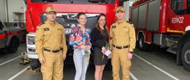 Komendantów oraz dwie kobity (pracownice cywilne), którzy stoją przed wozem strażackim i pozują do zdjęcia. Kobiety trzymają w rękach kopertę oraz różę.