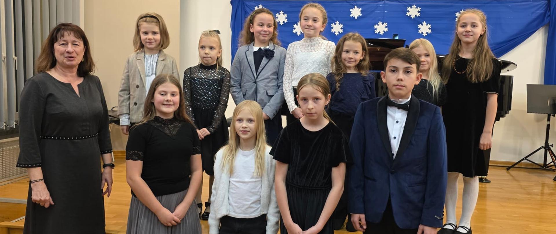 Na zdjęciu odświętnie ubrani uczniowie, którzy są ustawieni w dwóch rzędach. Zdjęcie zostało wykonane podczas koncertu uczniów klasy fortepianu Pani Katarzyny Olszewskiej. Po lewej stronie obrazka kobieta brunetka w czarnej sukience. W tle niebieski materiał z białym śnieżynkami. Podłoga wykonana z brązowego parkietu.