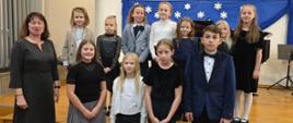 Na zdjęciu odświętnie ubrani uczniowie, którzy są ustawieni w dwóch rzędach. Zdjęcie zostało wykonane podczas koncertu uczniów klasy fortepianu Pani Katarzyny Olszewskiej. Po lewej stronie obrazka kobieta brunetka w czarnej sukience. W tle niebieski materiał z białym śnieżynkami. Podłoga wykonana z brązowego parkietu.