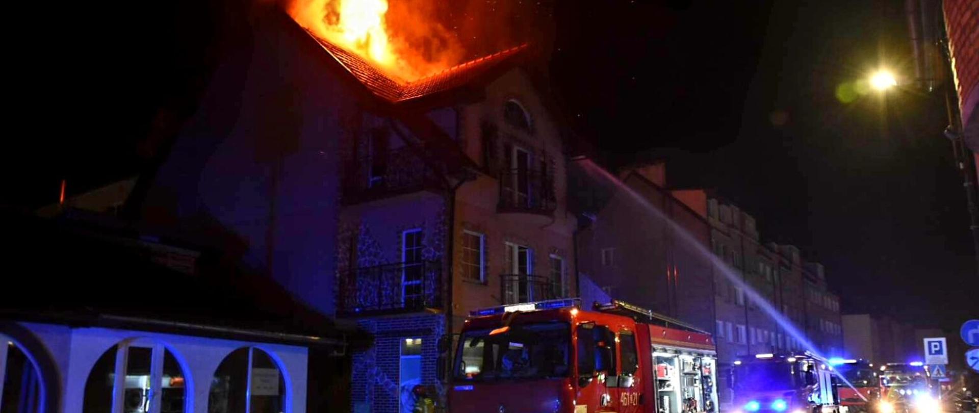 Zdjęcie przedstawia pożar budynku mieszkalnego w porze nocnej oraz wozy strażackie które biorą udział w akcji.
