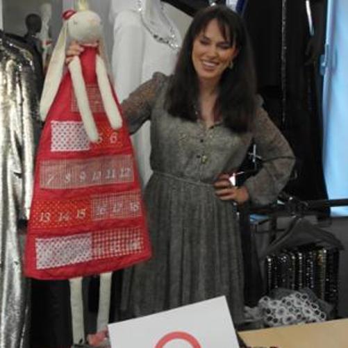 A women is presenting a doll, behind the woman there are clothes 