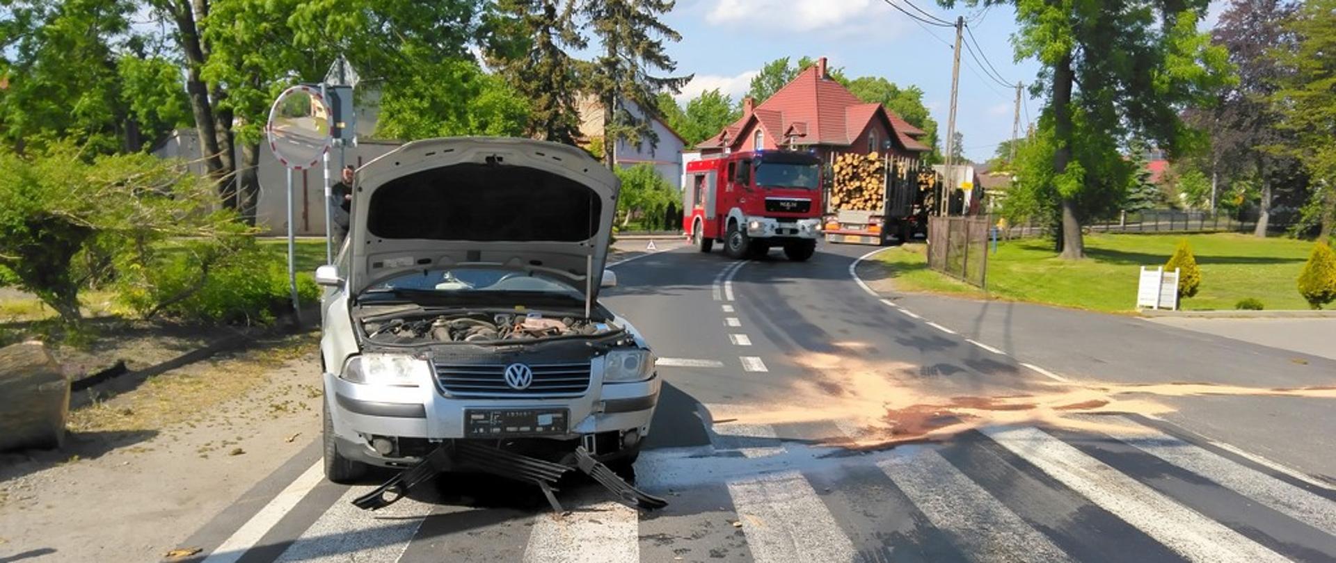 Dzień. Widok na drogę o powierzchni asfaltowej. Samochód osobowy marki Volkswagen w kolorze srebrnym, skierowany przodem, stoi częściowo na lewym pasie drogi a częściowo na poboczu szutrowym. Samochód przednimi kołami stoi na przejściu dla pieszych. Podniesiona maska pojazdu. Widoczne zniszczenia dolnej części przedniego zderzaka. Z prawej strony pojazdu widać ciemne plamy na asfalcie częściowo pokryte materiałem sypkim (sorbentem) w kolorze łososiowym. W tle samochód ratowniczo-gaśniczy w kolorze czerwonym blokuje przejazd drogą. Za nim stoi samochód ciężarowy z przyczepą załadowanym belkami drewna.. W tel widać budynki mieszkalne. 