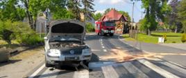 Dzień. Widok na drogę o powierzchni asfaltowej. Samochód osobowy marki Volkswagen w kolorze srebrnym, skierowany przodem, stoi częściowo na lewym pasie drogi a częściowo na poboczu szutrowym. Samochód przednimi kołami stoi na przejściu dla pieszych. Podniesiona maska pojazdu. Widoczne zniszczenia dolnej części przedniego zderzaka. Z prawej strony pojazdu widać ciemne plamy na asfalcie częściowo pokryte materiałem sypkim (sorbentem) w kolorze łososiowym. W tle samochód ratowniczo-gaśniczy w kolorze czerwonym blokuje przejazd drogą. Za nim stoi samochód ciężarowy z przyczepą załadowanym belkami drewna.. W tel widać budynki mieszkalne. 