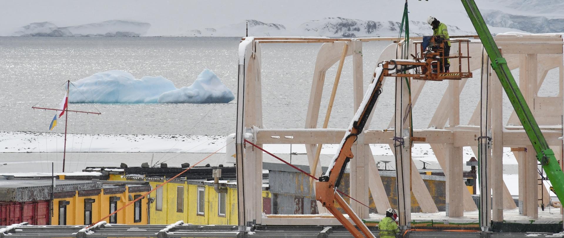 Widok budowy dużej konstrukcji z drewna, na podnośniku stoi osoba w żółtej kurtce, w tle z lewej żółty parterowy budynek stacji, dalej morze na którym pływa mała góra lodowa.