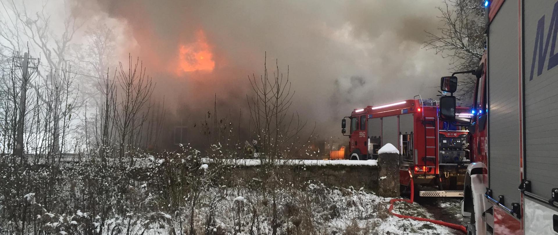 Zdjęcie przedstawia pożar budynku wielorodzinnego stojącego w płomieniach, po prawej stronie znajdują się wozy strażackie.