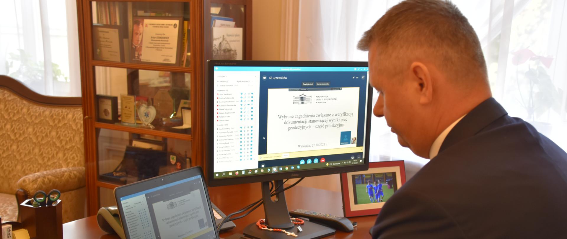 Na zdjęciu znajduje się mężczyzna siedzący za biurkiem przed dwoma komputerami