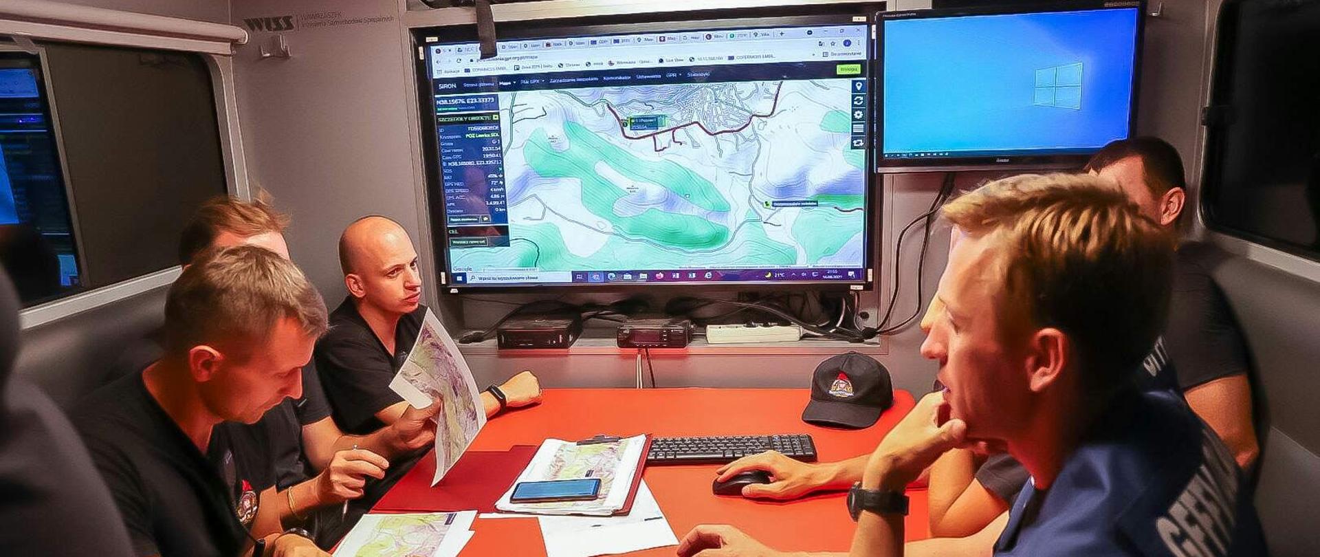Dowództwo polskiej misji analizuje sytuację pożarową w samochodzie dowodzenia i łączności. W tle mapy terenu, na którym działają strażacy.