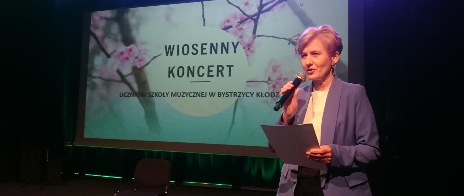 Zdjęcie przedstawia wicedyrektor zapowiadającą uczestników w trakcie majowego koncertu wiosennego w MGOK w Bystrzycy Kłodzkiej. 
