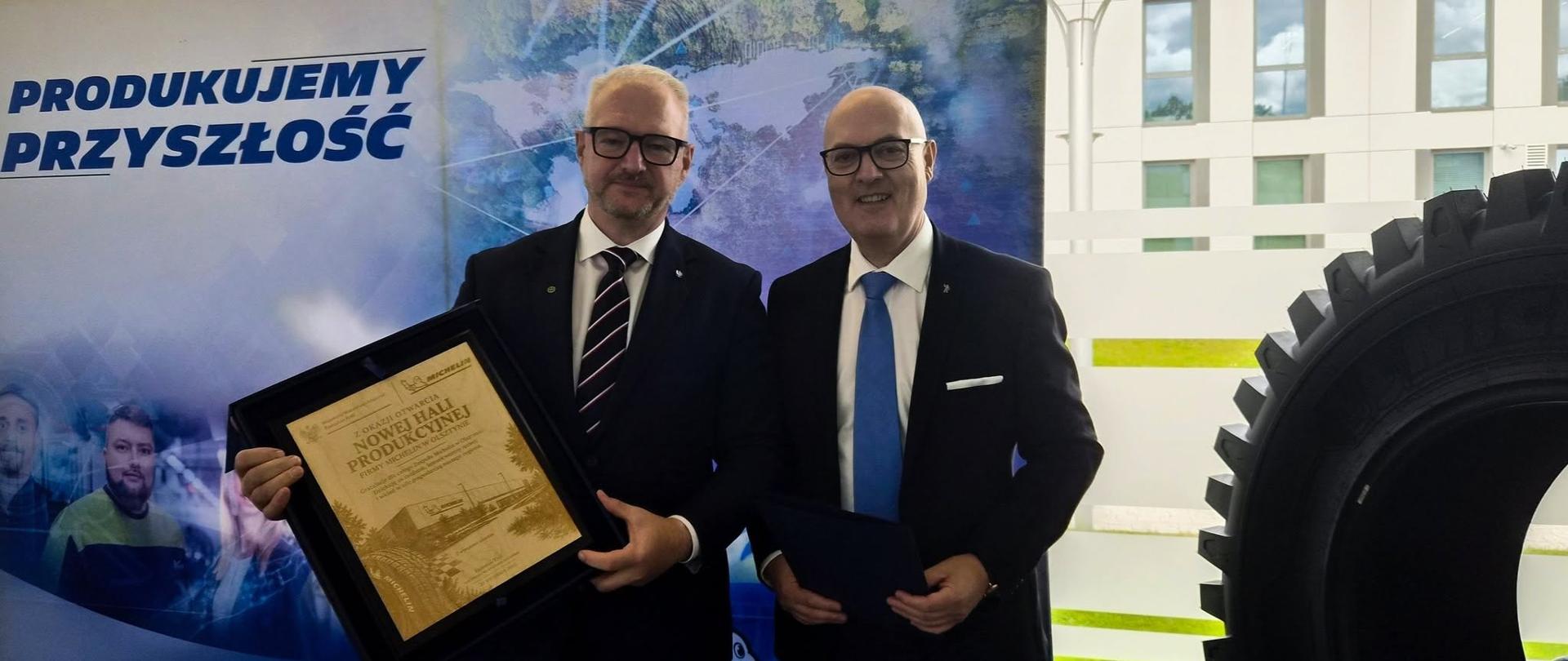 Mężczyzna w garniturze daje drugiemu mężczyźnie w garniturze pamiątkową tabliczkę. 
