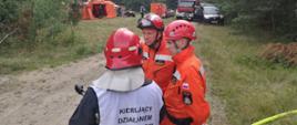 Kierujący działaniem ratowniczym przekazuje strażakom z grupy poszukiwawczej zadania, które mają wykonać. Za nimi znajduje się obozowisko.