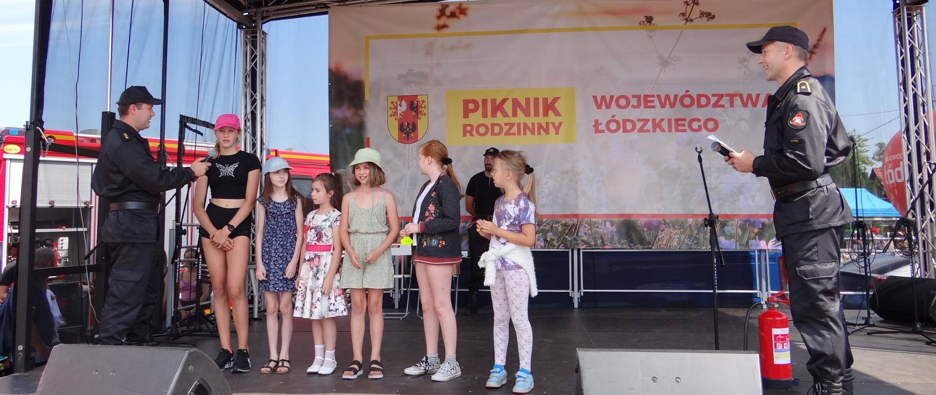 Na scenie stoją dzieci. Dwóch strażaków w mundurach przeprowadza konkurs wiedzy o pożarnictwie. Na scenie stoją kolumny głośnikowe, statywy do mikrofonów i gaśnica. Jest pogodnie.