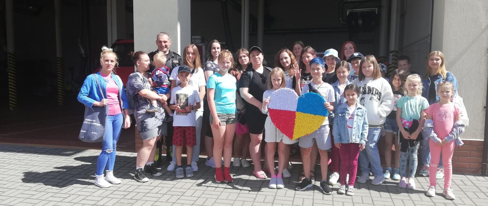 Przed bramami garażowymi Jednostki Ratowniczo-Gaśniczej w Człuchowie stoi grupa dzieci wraz ze swoimi opiekunami. Z grupą stoi Dowódca Jednostki. Z przodu dzieci trzymają duże cerce w kolorach flag państwowych Polski i Ukrainy.