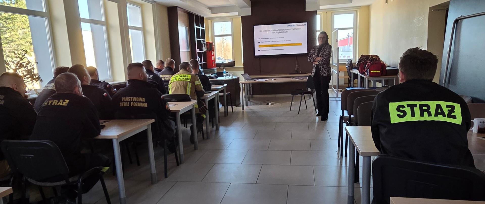 Strażacy PSP uczestniczą w szkoleniu prowadzonym w sali dydaktycznej, słuchając prezentacji wyświetlanej na ekranie.