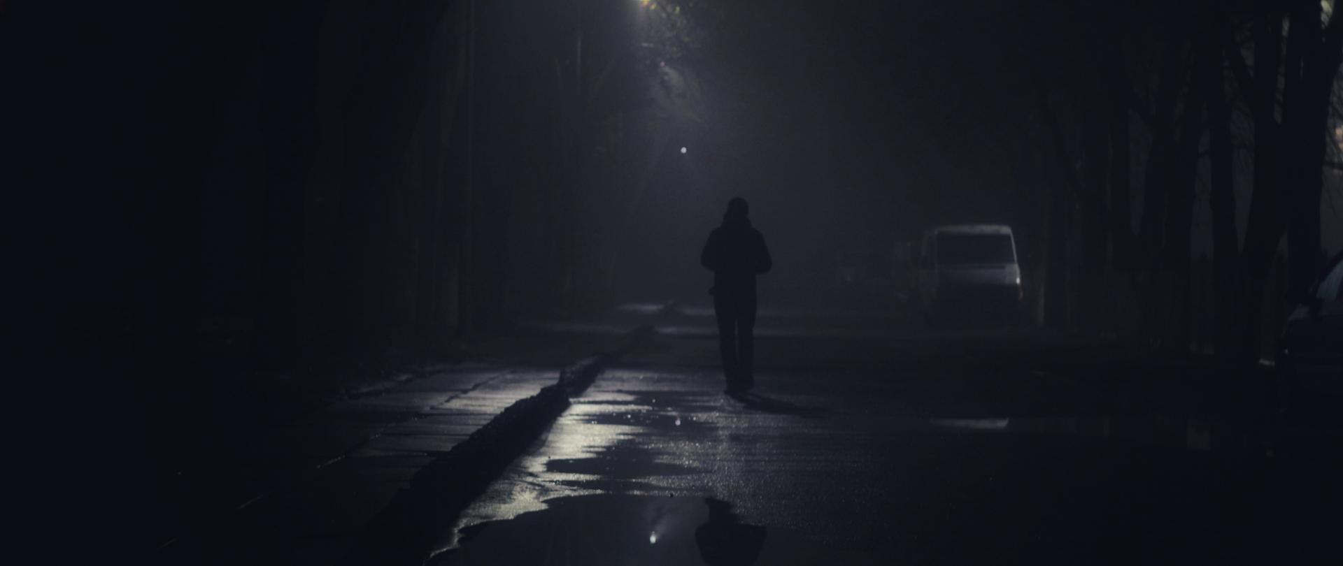 Ciemna ulica i sączące się światło latarni. Ulicą idzie człowiek ubrany na ciemno, ledwo widoczny na drodze.