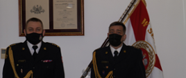 dwóch strażaków w ciemnych mundurach ze złotymi sznurami, w białych koszulach i czarnych krawatach, z maseczkami ochronnymi na twarzach stoi na baczność, w tle sztandar