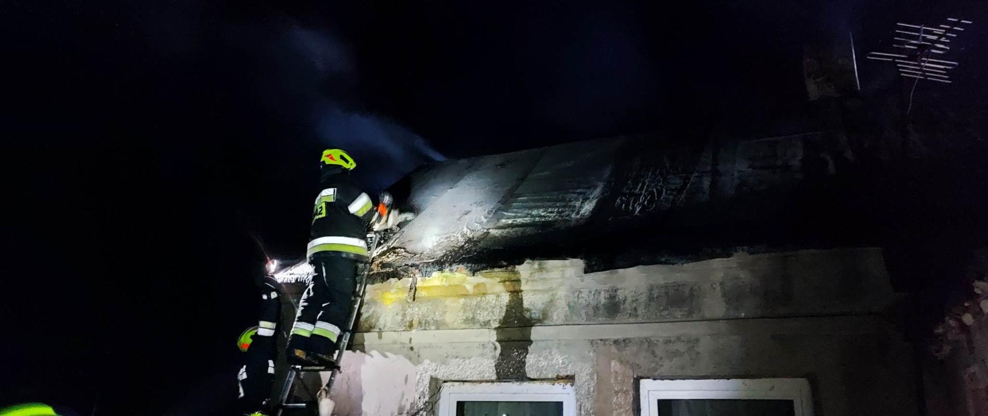 Na zdjęciu częściowo widać parterowy murowany budynek mieszkalny, w którym doszło do pożaru. Widać strażaka w ciemnym stroju, który stoi na drabinie opartej o ścianę budynku. Obok budynku stoi drugi strażak, który świeci latarką. Jest ciemno. 