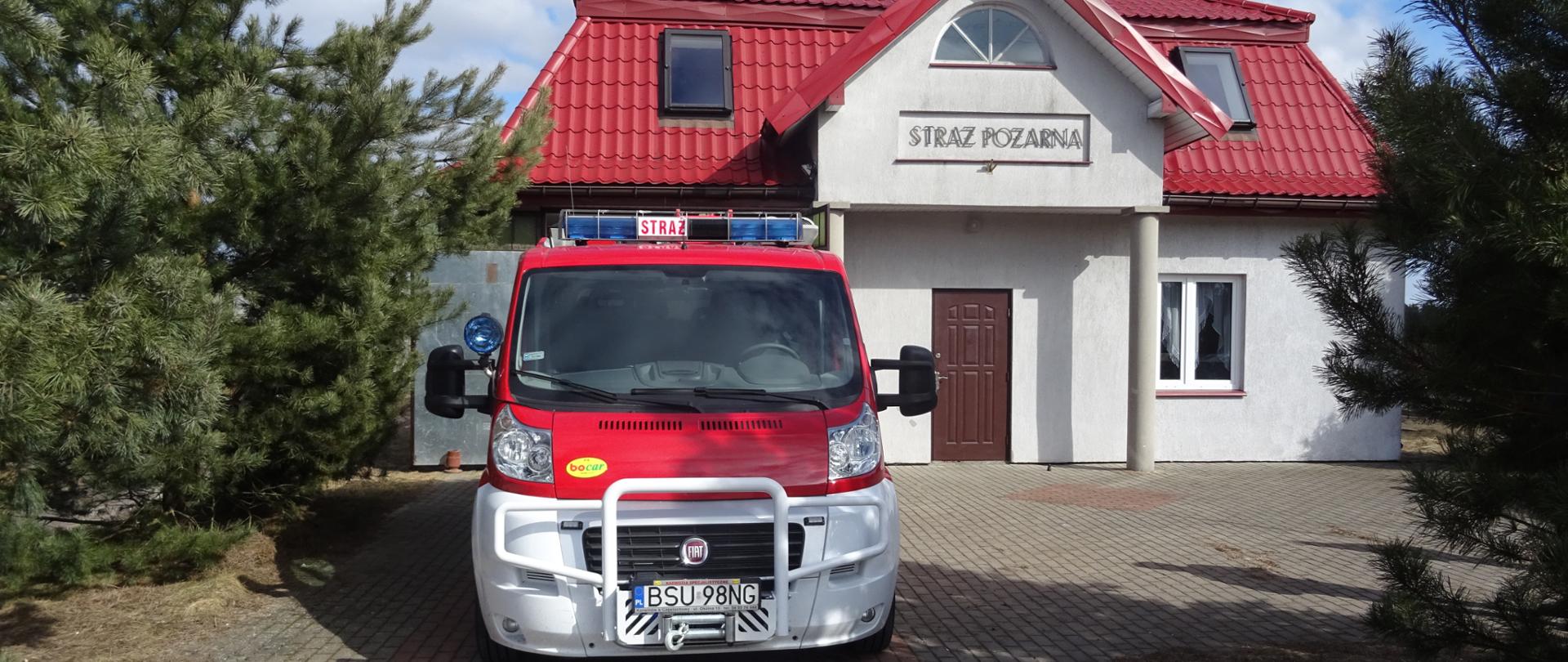 Zdjęcie przedstawia samochód pożarniczy w tle budynek remizy OSP Sucha Wieś budynek o jasnych ścianach spadzistym dachem w kolorze czerwonym napis na elewacji straż pożarna po bokach budynku rosną drzewa