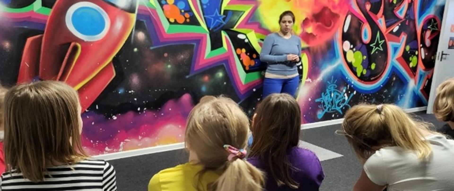 Przedstawicielka Sanepidu w Gdańsku stoi zwrócona w stronę dzieci siedzących na krzesłach. W tle znajduje się kolorowe graffiti. Kobieta rozmawia z dziećmi na temat bezpiecznych ferii zimowych.
