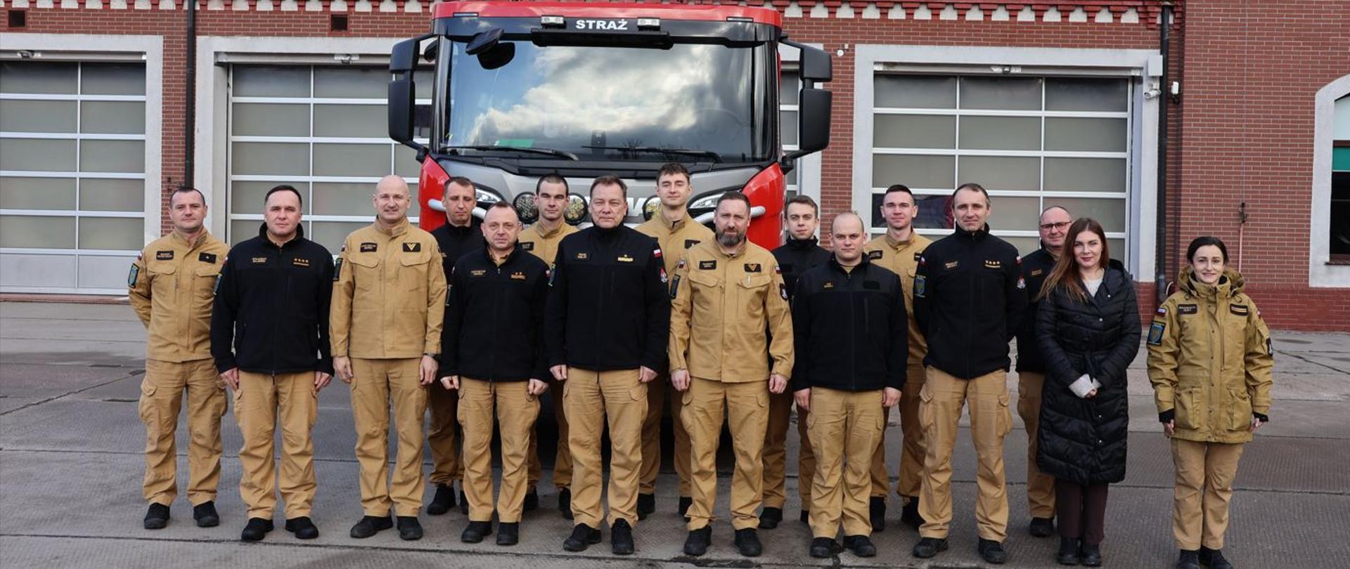 Grupa strażaków w mundurach i pracowników stoi w szeregu przed remizą strażacką, a za nimi zaparkowany jest czerwony wóz strażacki