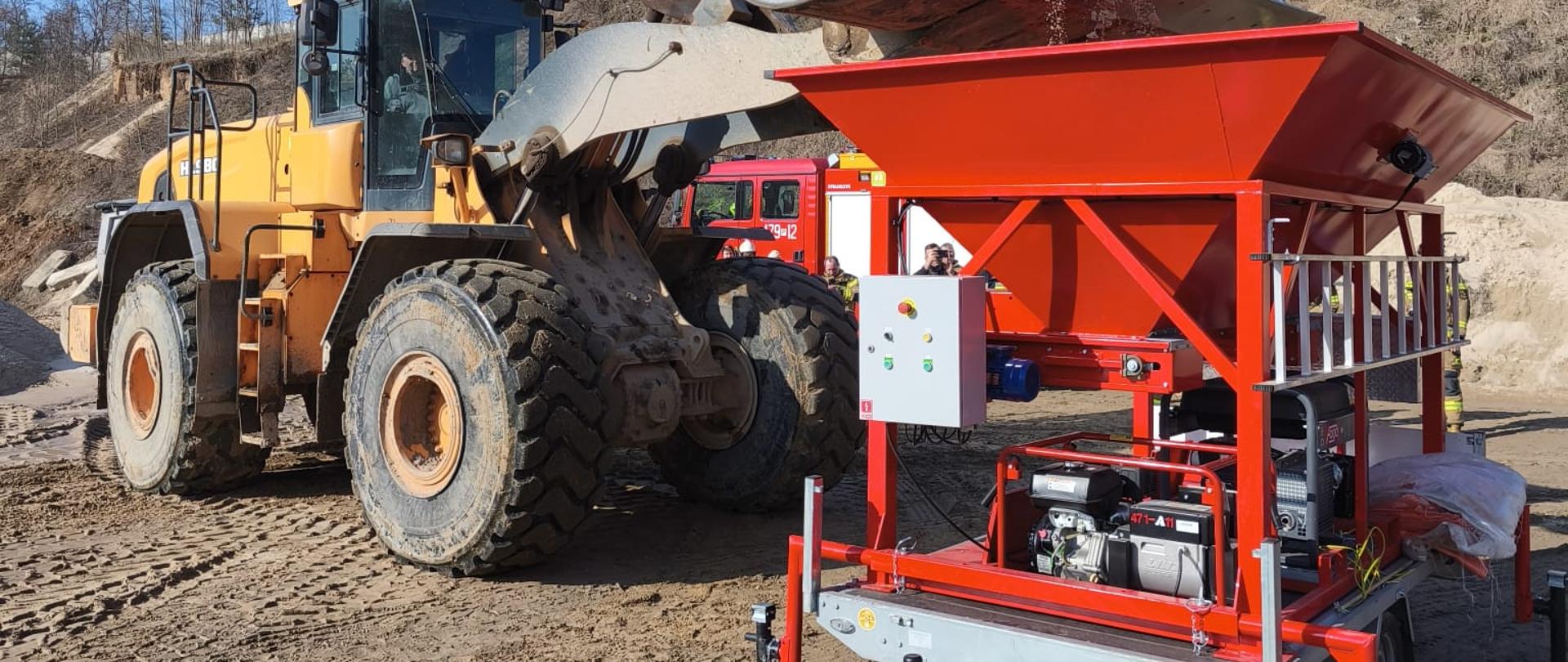 Ładowarka kołowa wysypuje piasek do dużego czerwonego zasobnika mobilnej maszyny do napełniania worków ustawionej na przyczepie.