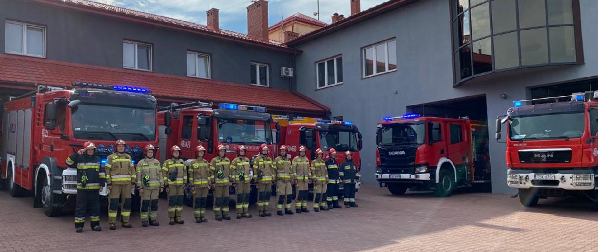 Na zdjęciu widać strażaków KP PSP Skarżysko-Kamienna ubranych w ubrania bojowe, oddających hołd Powstańcom Warszawskim. Stoją oni przed garażami, są wyprowadzone również samochody bojowe z włączonymi sygnałami świetlnymi i dźwiękowymi. W tle widać budynek Komendy Powiatowej. 