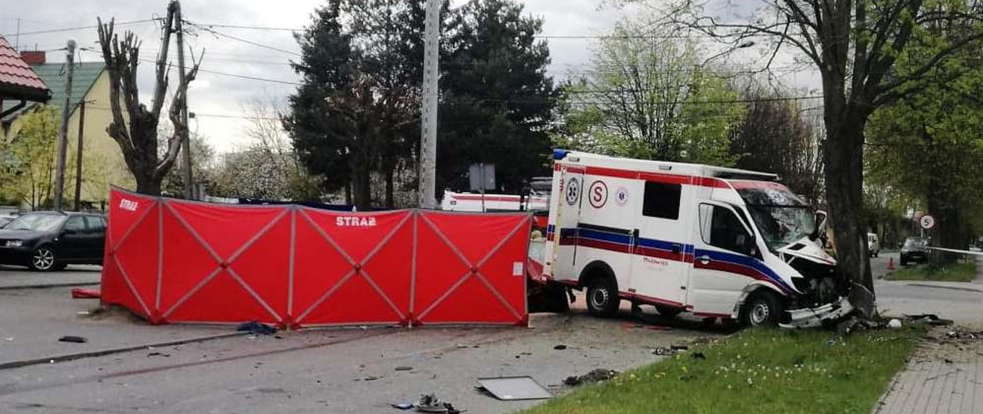 Wypadek w Ostrowi Mazowieckiej na ul. Szkolnej. Na zdjęciu rozbita karetka, która uderzyła w drzewo. Na jezdni rozstawiony jest parawan straży pożarnej.