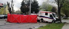 Wypadek w Ostrowi Mazowieckiej na ul. Szkolnej. Na zdjęciu rozbita karetka, która uderzyła w drzewo. Na jezdni rozstawiony jest parawan straży pożarnej.